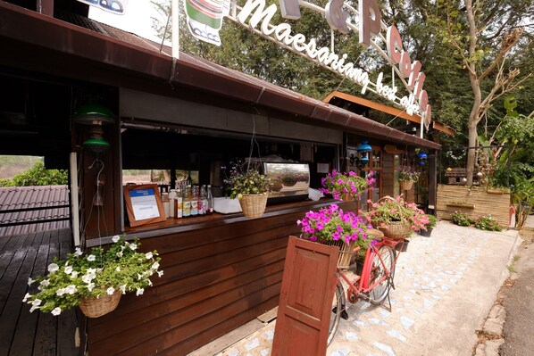 A cafe with a wooden exterior and a sign that reads "Maesananing her cafe." There are potted plants and a bicycle with baskets of flowers outside.