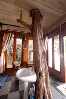 Un baño rústico con un gran tronco de árbol, un lavamanos blanco y una ventana que ofrece vistas al exterior.