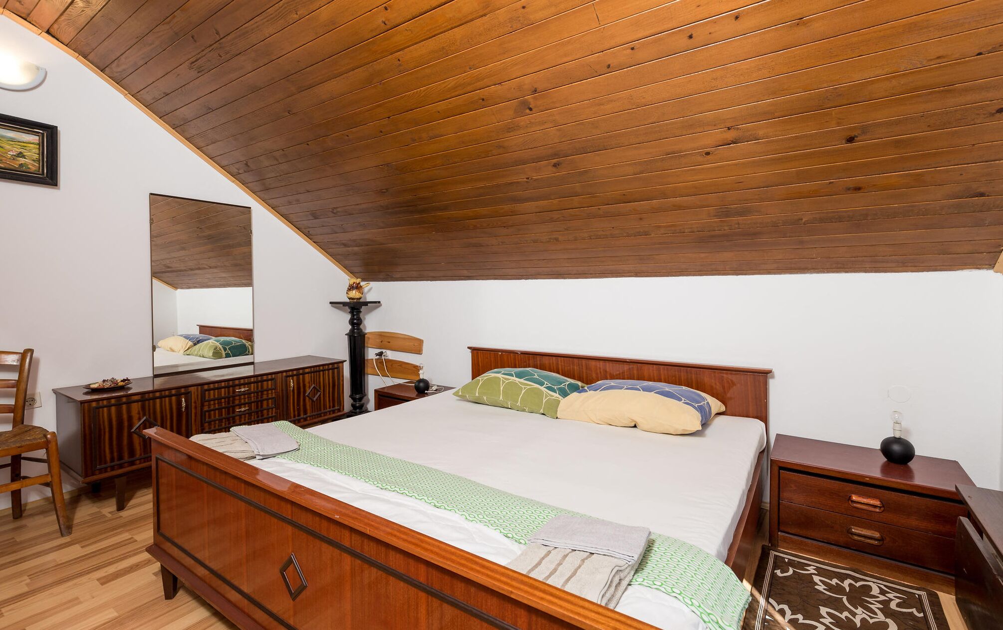 Une chambre avec un plafond en bois, un lit avec des oreillers verts et beiges, une commode en bois et une table de chevet avec une lampe.
