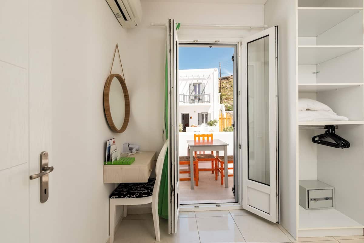 A room with a white door, a mirror, a desk with a chair, and a view of an outdoor area with a table and chairs.
