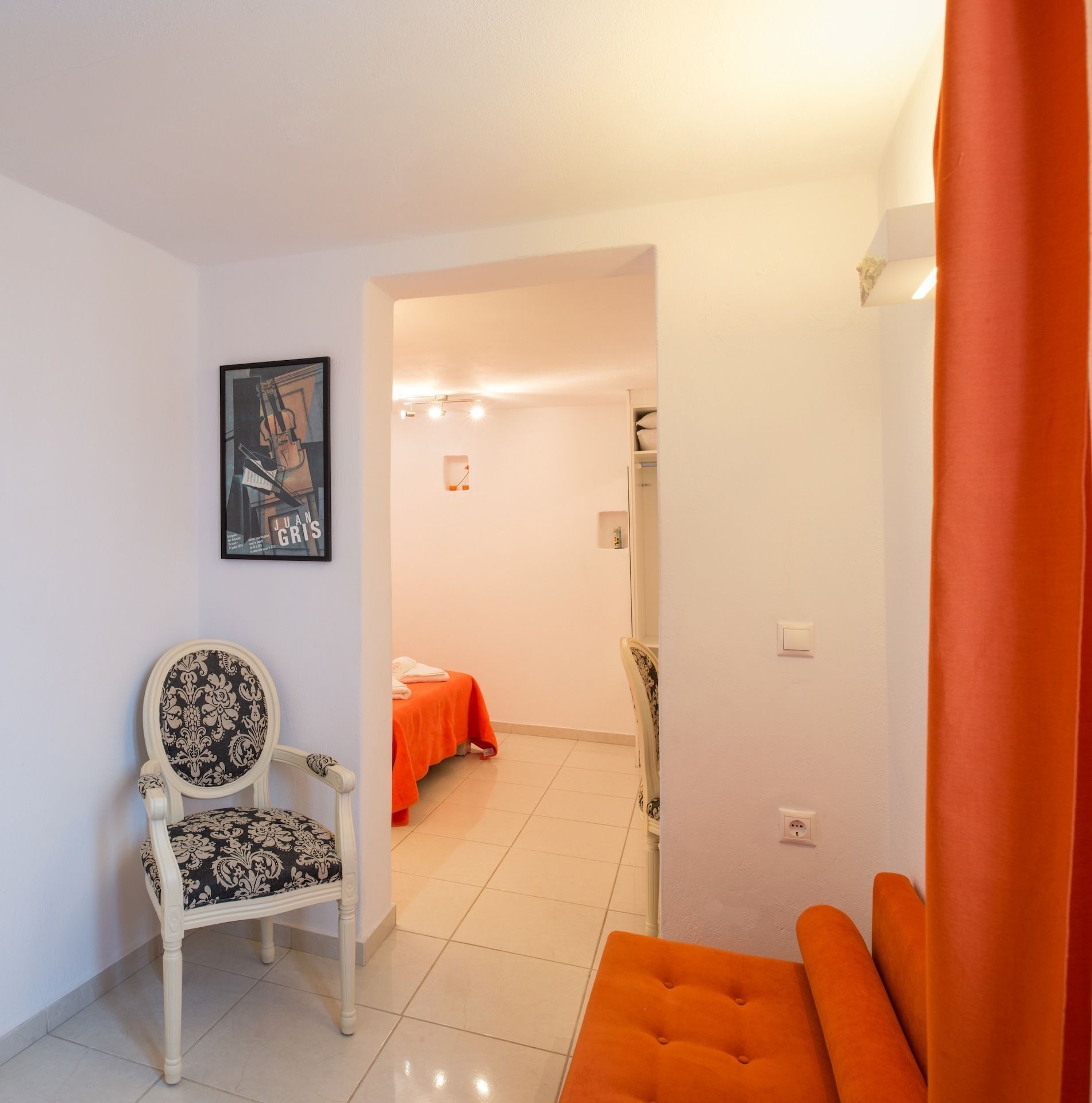 A room with a white chair, an orange ottoman, a bed with an orange bedspread, and a framed picture on the wall.