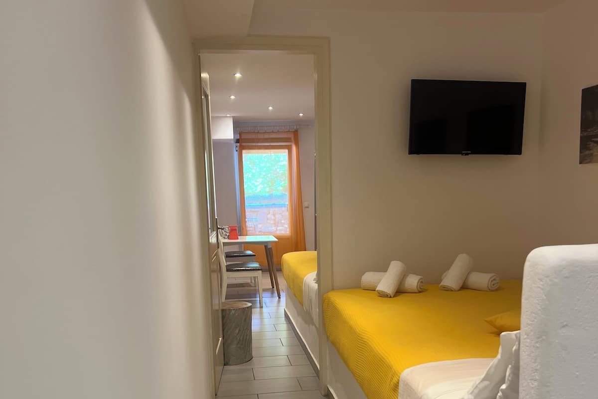 A hotel room with two beds, one with a yellow bedspread and the other with a white bedspread. There is a television mounted on the wall, a window with curtains, and a small table with a chair.