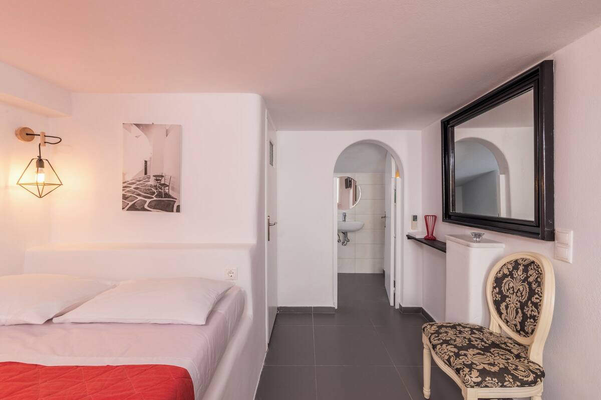 A bedroom with a bed, a chair, a mirror, and a bathroom visible through an open door.