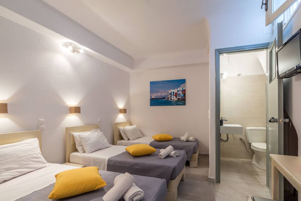 A hotel room with three beds, each with a pillow and a rolled towel. There is a painting on the wall and a bathroom visible through an open door.