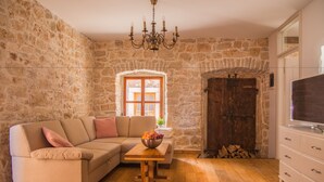 Un salón con pared de piedra, suelo de madera, sofá color beige, mesa de madera con frutero, televisor sobre un mueble blanco y una puerta grande de madera.