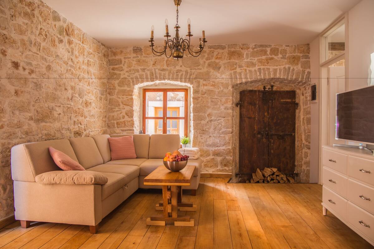 Un salón con pared de piedra, suelo de madera, sofá color beige, mesa de madera con frutero, televisor sobre un mueble blanco y una puerta grande de madera.