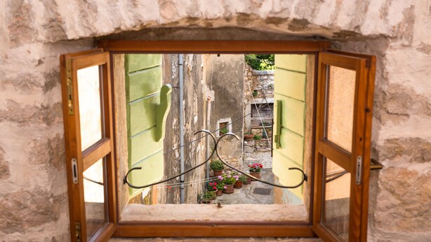Una ventana abierta con vista a una calle estrecha, edificios y plantas en macetas.