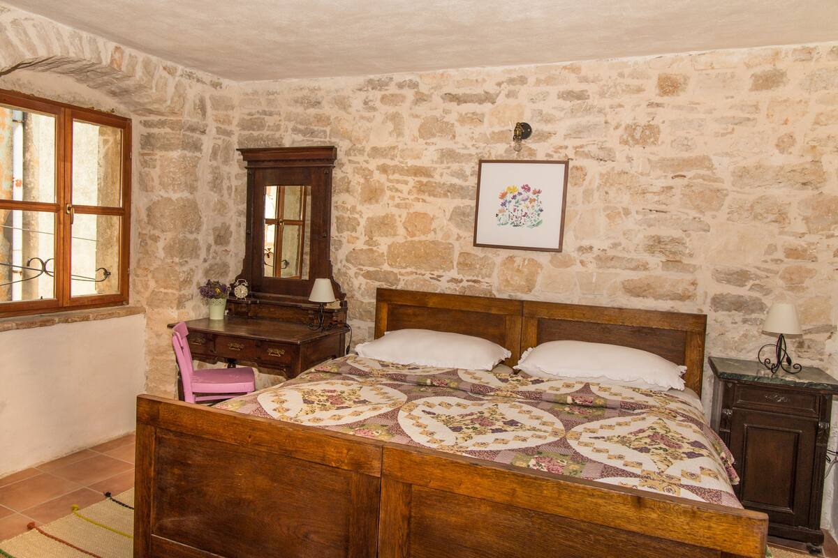 Un dormitorio con una cama de madera, pared de piedra, cómoda de madera y ventana con cortinas.