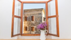 Una habitación con ventana abierta que da a un edificio de piedra con contraventanas de madera. Un jarro blanco con flores moradas está sobre el alféizar.