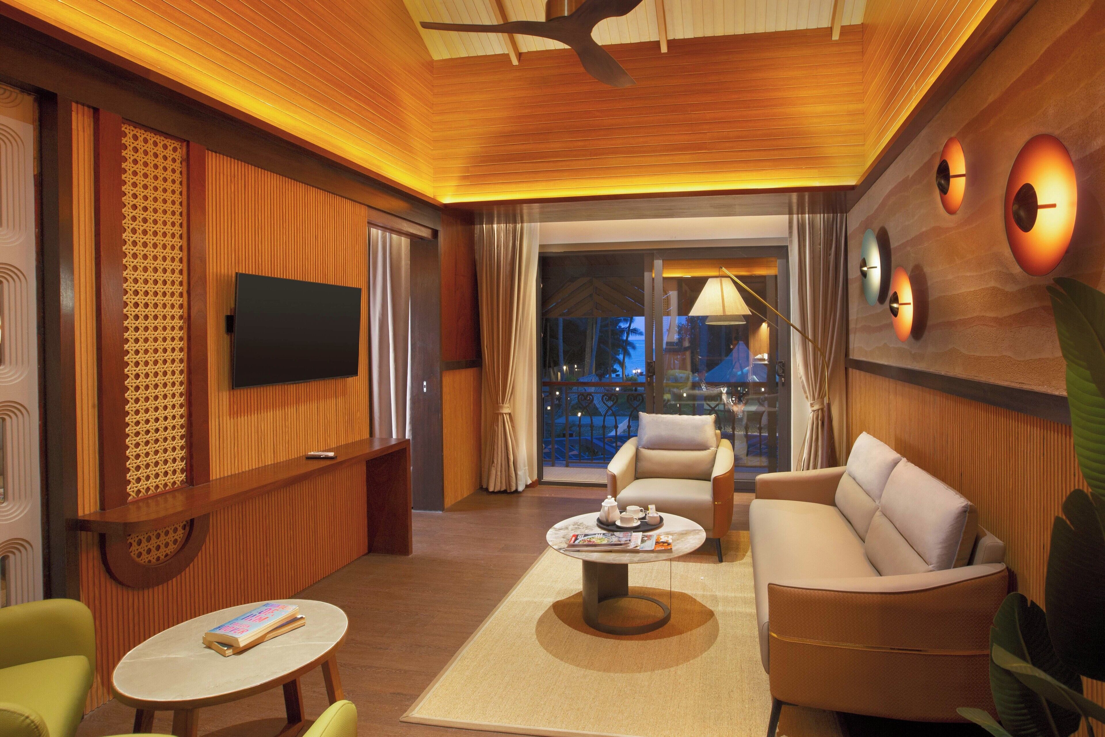 A modern hotel room with a flat-screen TV, a sofa, a coffee table, and a view of the outdoors.