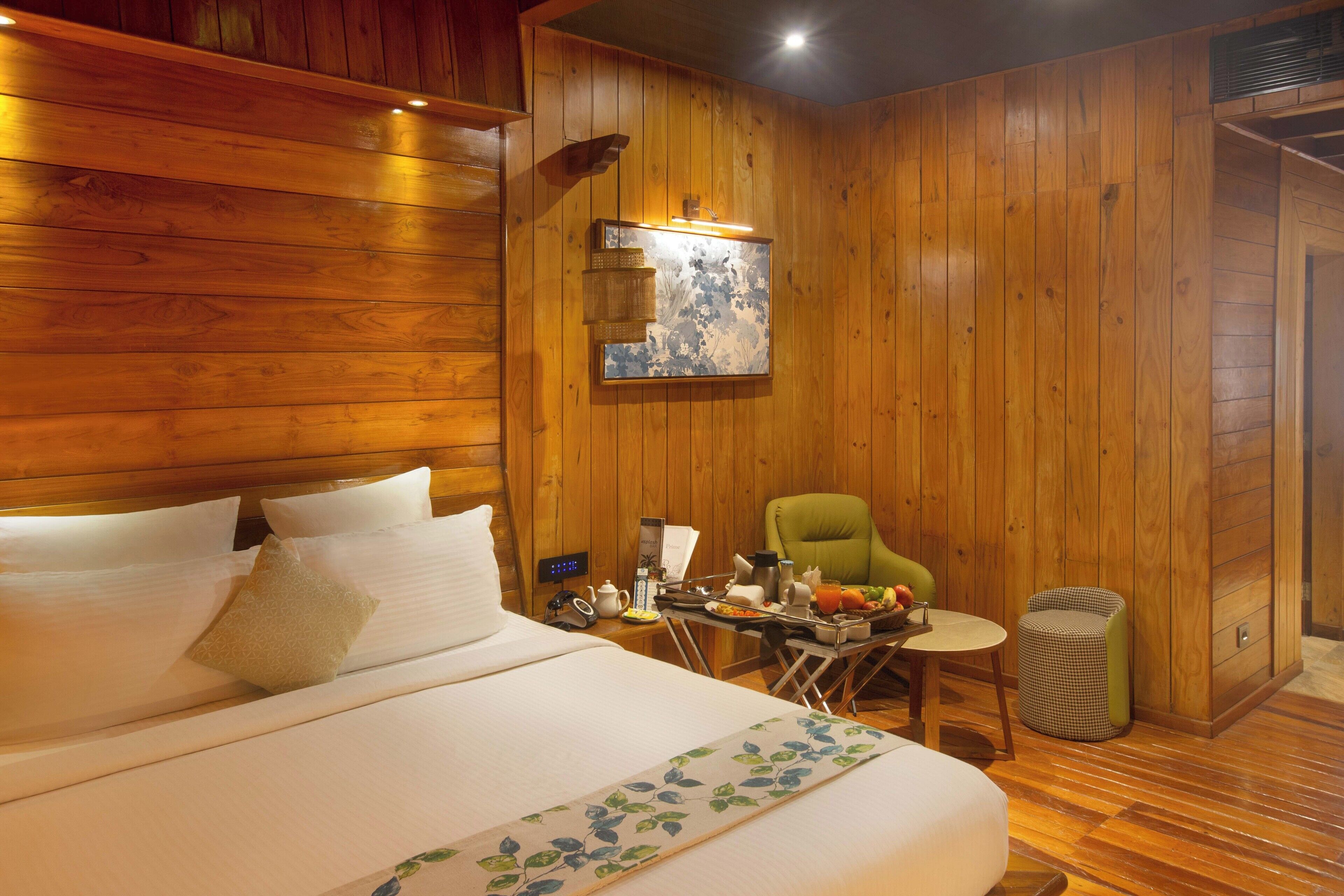 A wooden-paneled room with a bed, a small table with breakfast items, a chair, and a waste bin.