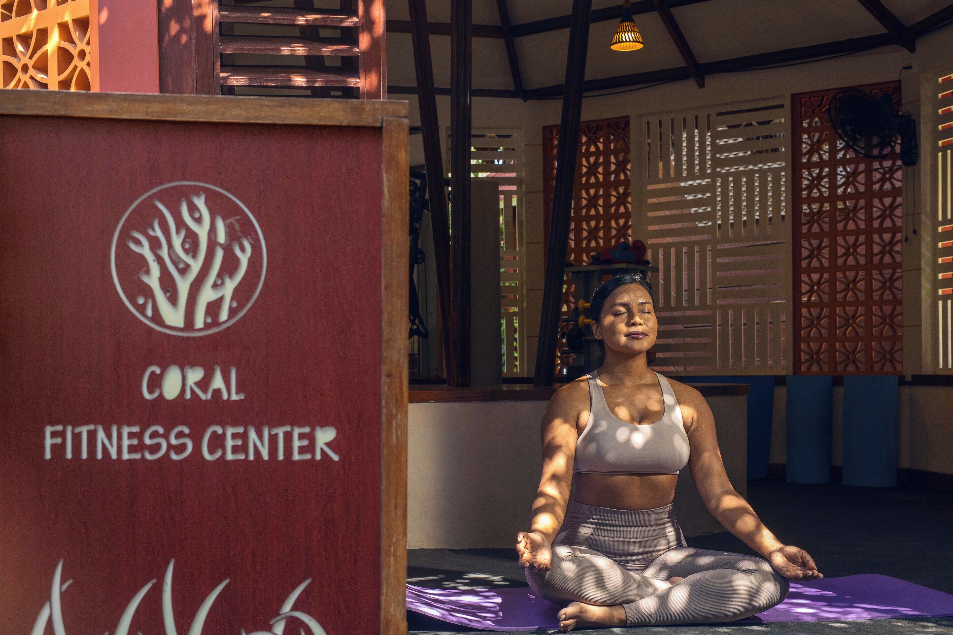 A woman meditating in a fitness center with a wooden structure labeled "CORAL FITNESS CENTER" in the background.