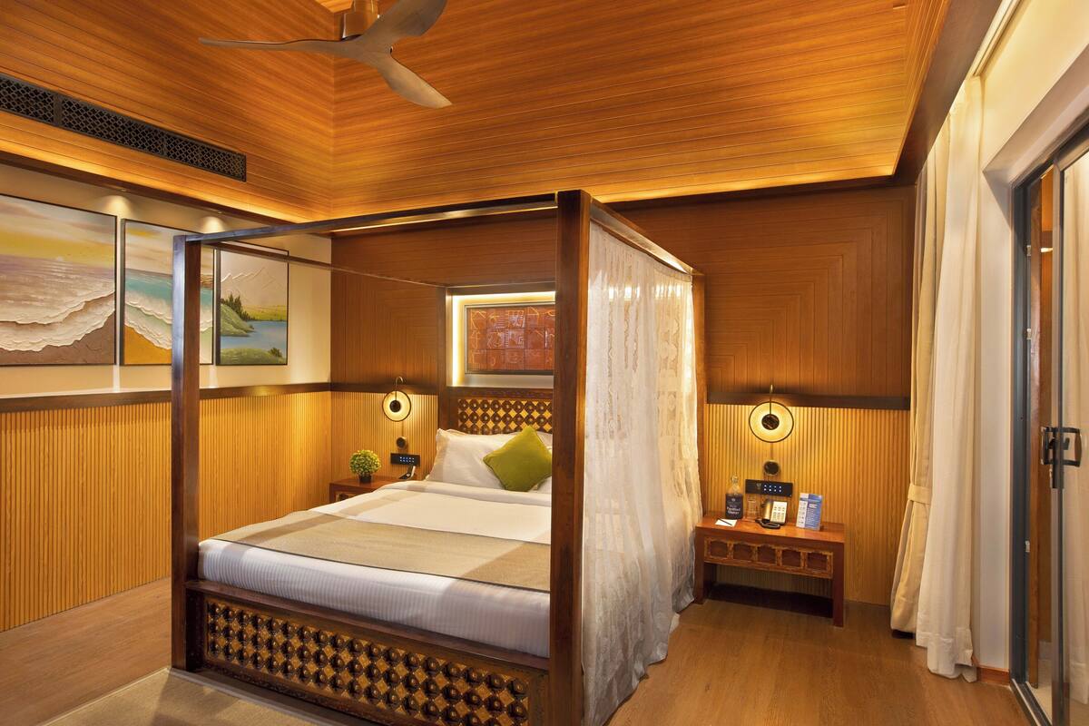A four-poster bed with a canopy, wooden paneling, a ceiling fan, a bedside table with a phone and a bottle, and a painting on the wall.