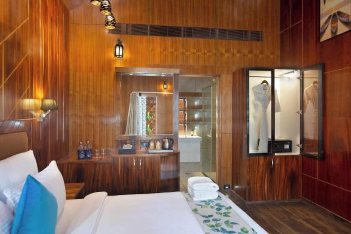 A hotel room with wooden paneling, a bed with white linens, a bedside table with toiletries, a mirror, and a shower area visible through a glass door.