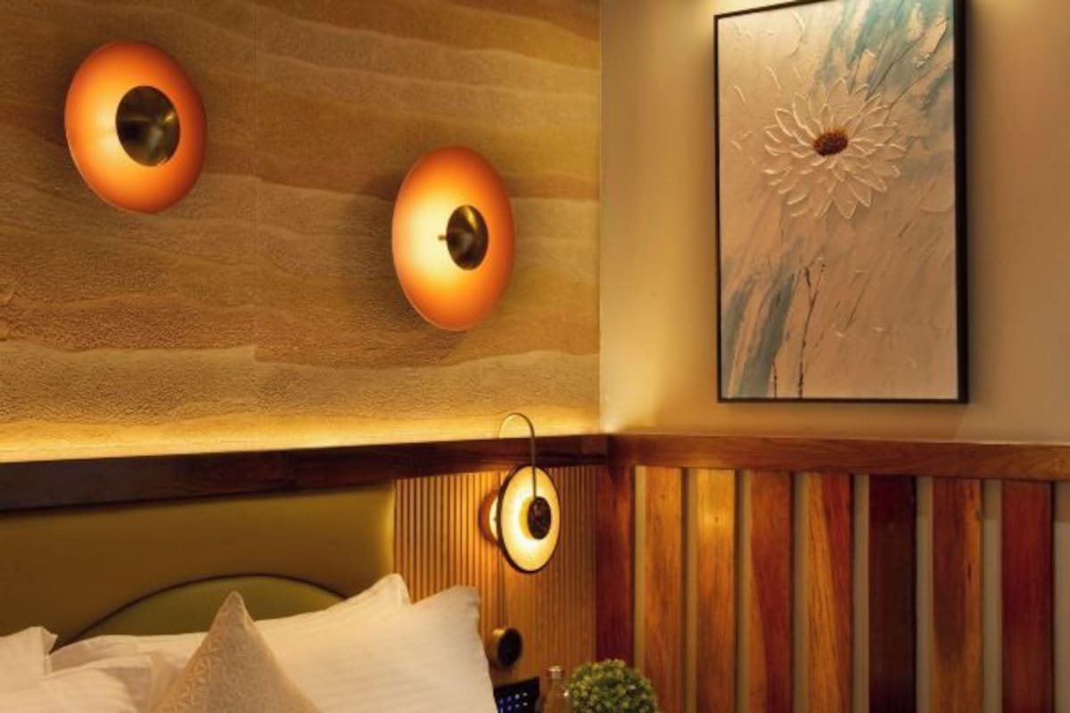 A hotel room with a wooden headboard, two wall-mounted lamps, a framed artwork, and a bed with pillows.