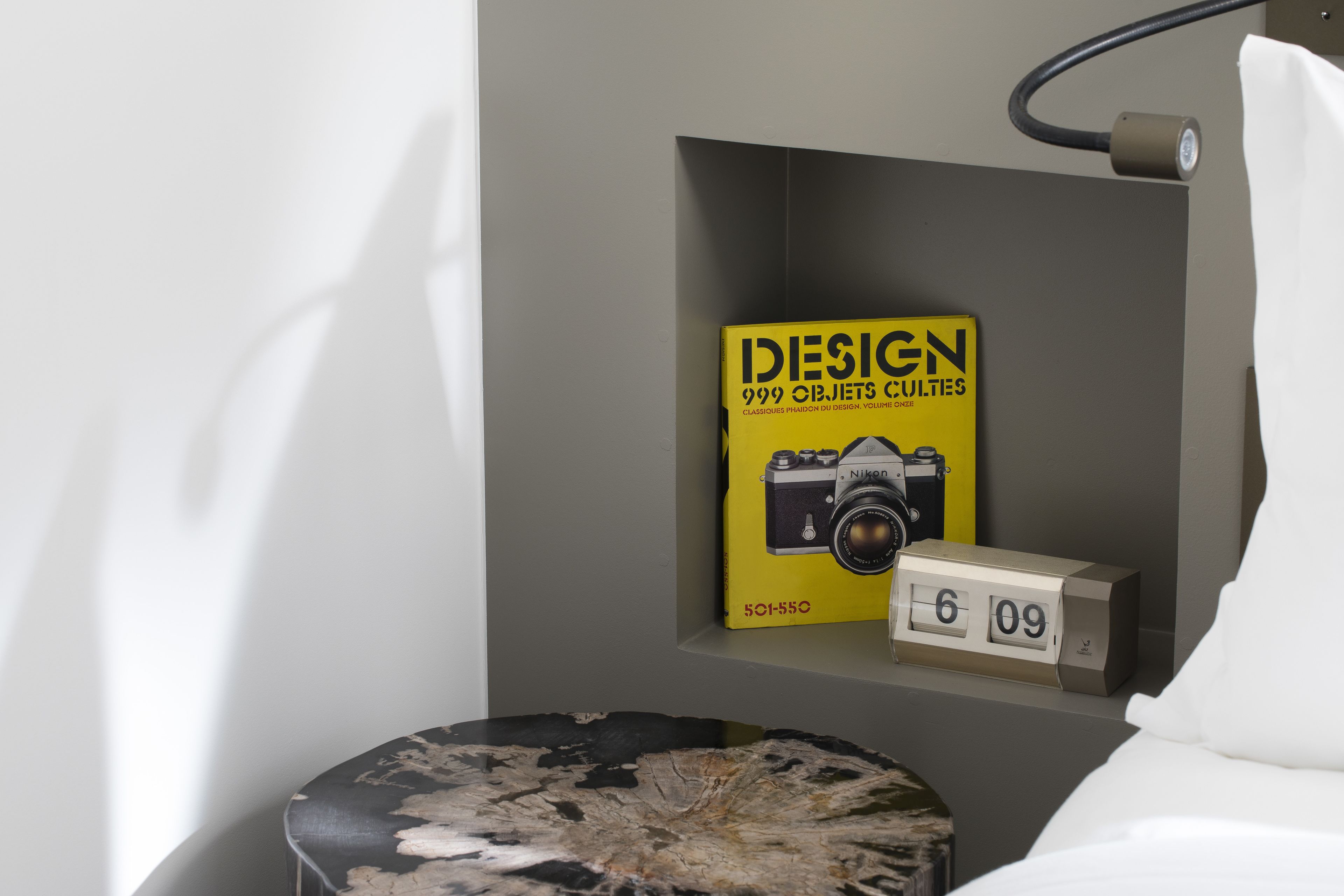 Un lit avec une literie blanche, une table de chevet avec une horloge et une étagère avec un livre et un appareil photo.