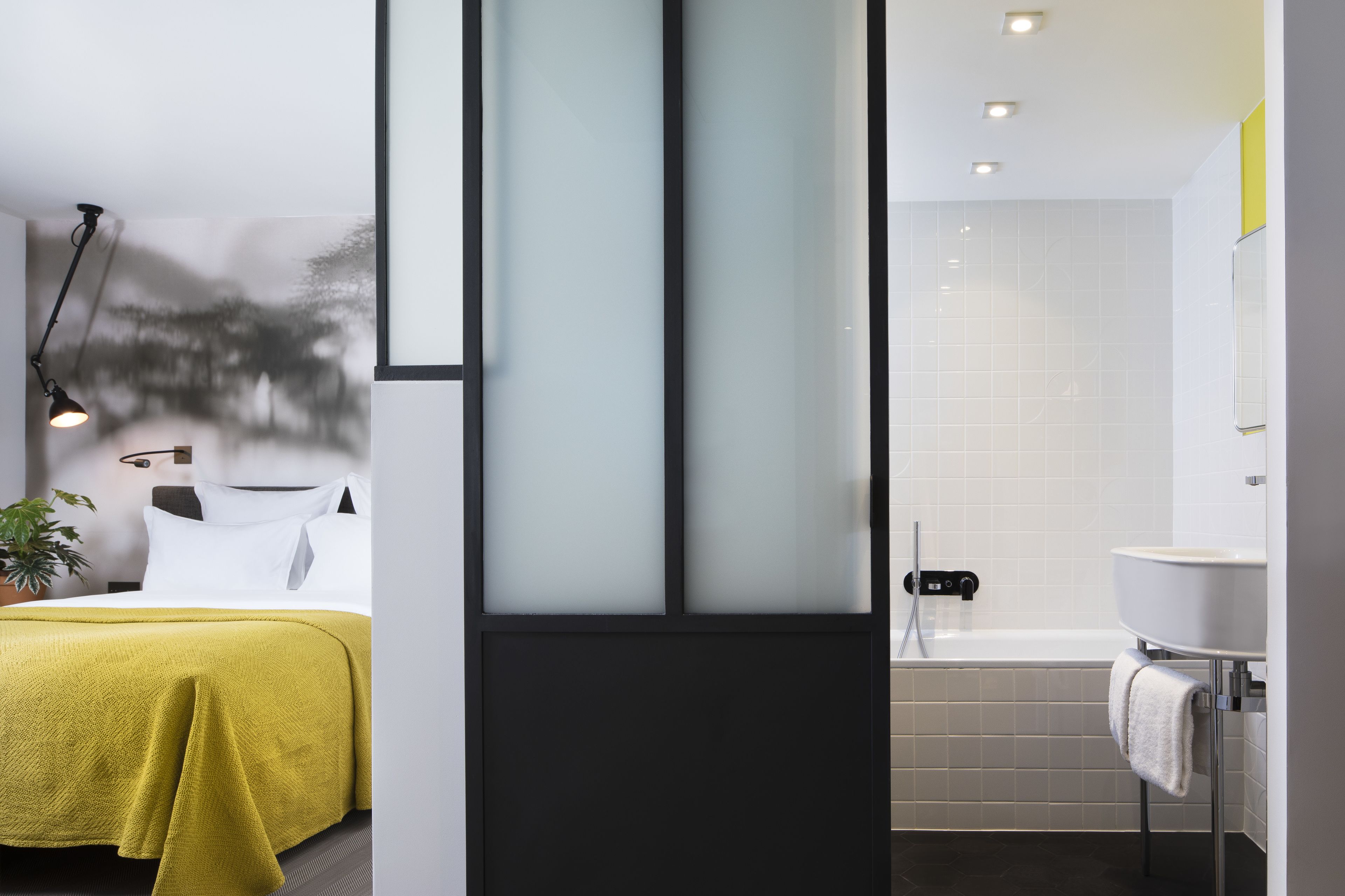 Una habitación de hotel moderna con una cama, una mesita de noche, una lámpara y un baño con bañera y lavamanos.