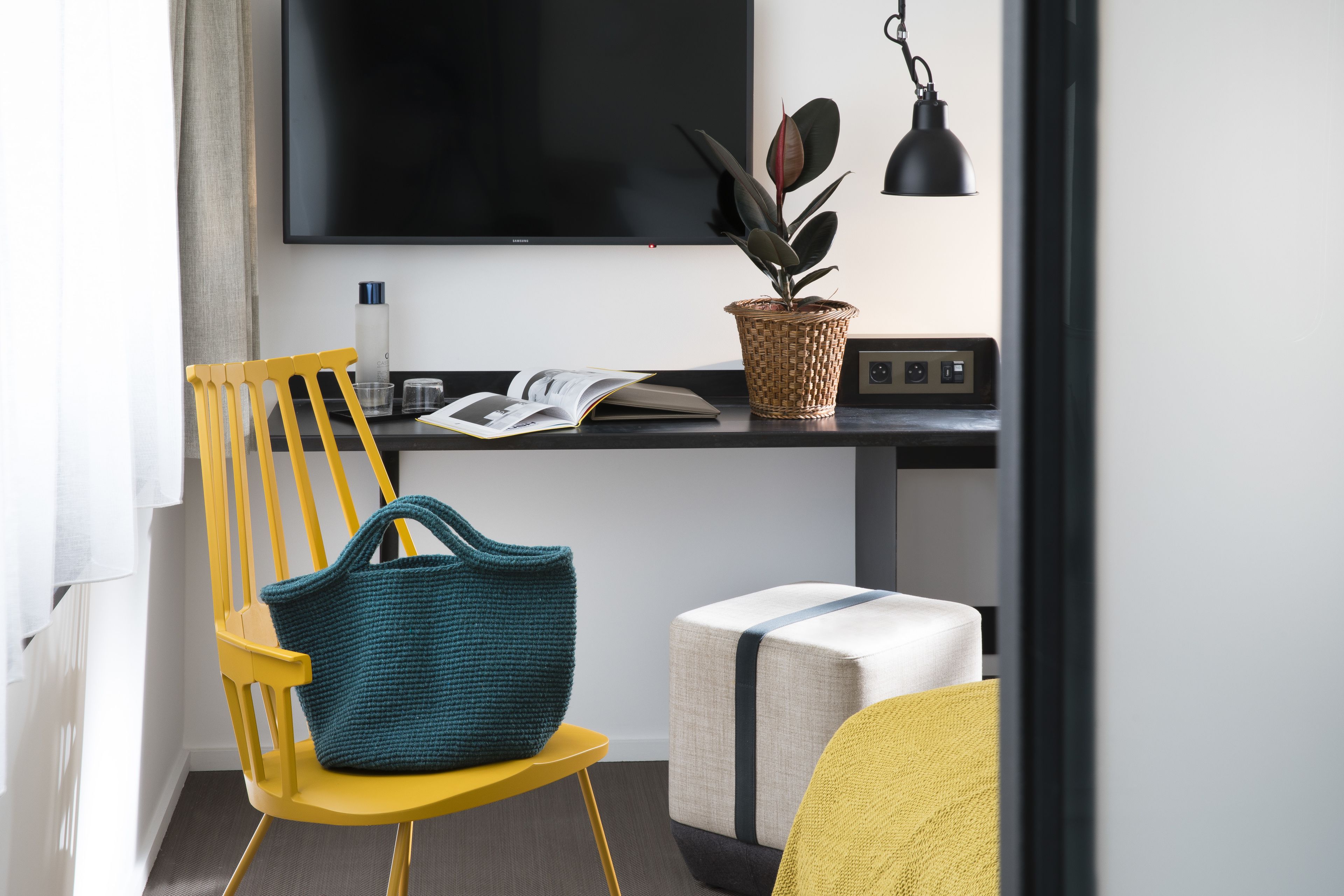 Quarto moderno com cadeira amarela, bolsa azul-turquesa, pequena mesa com uma planta, um livro e uma TV.
