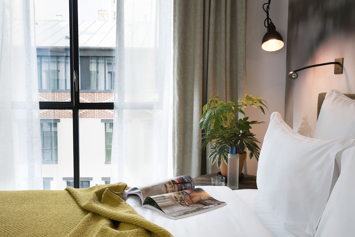 Una habitación de hotel con cama, una revista, una planta y un ventanal con cortinas.