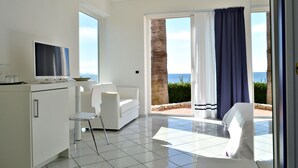A modern room with a TV, white furniture, a balcony with a view of the sea, and a tiled floor.