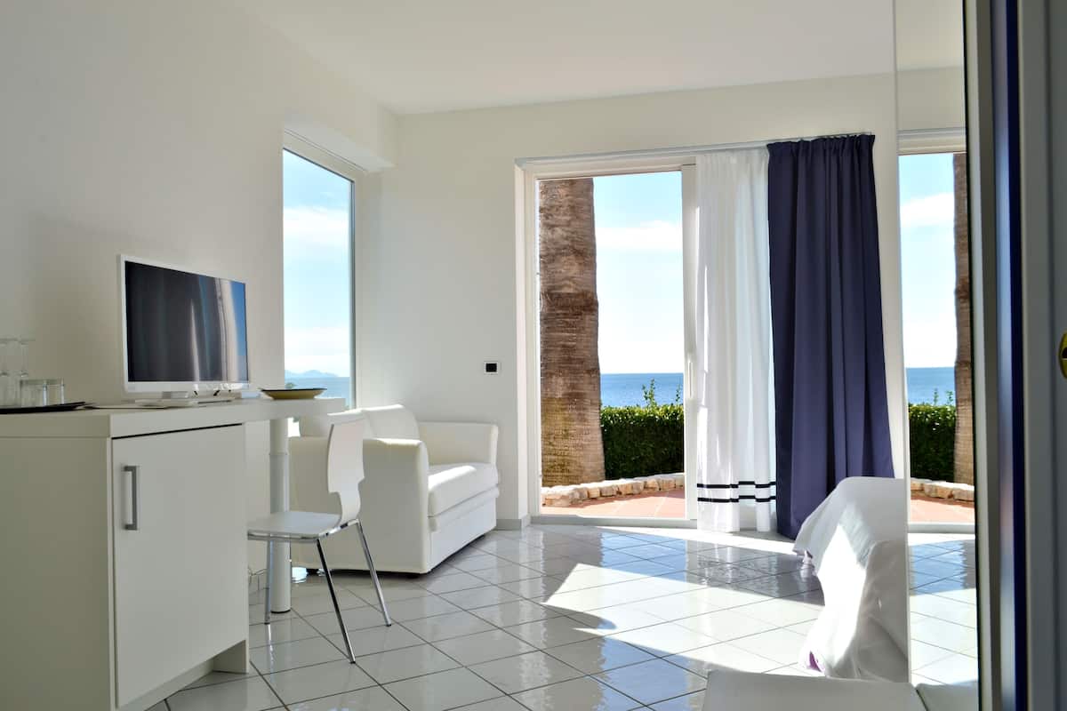 A modern room with a TV, white furniture, a balcony with a view of the sea, and a tiled floor.