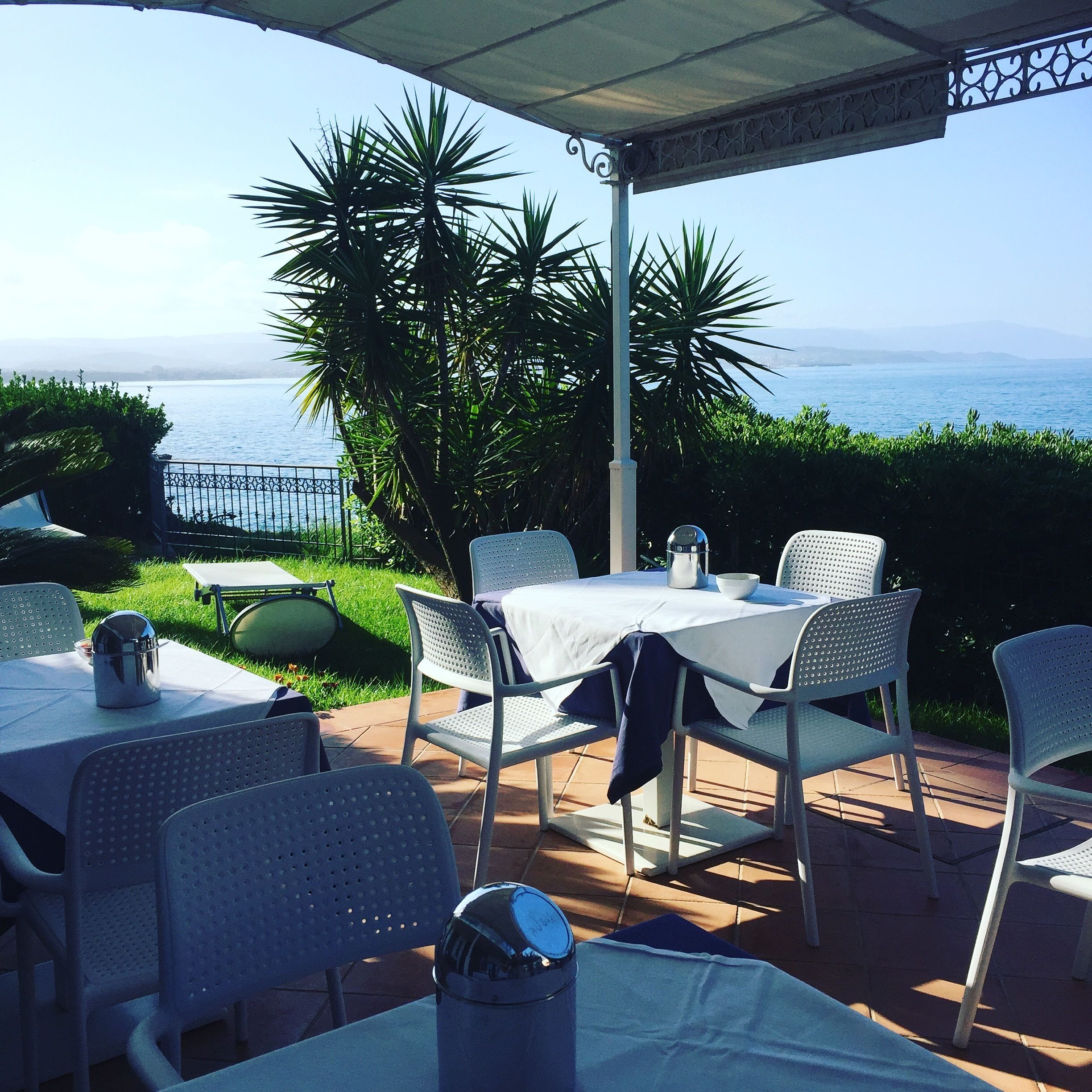 Un patio al aire libre con sillas y mesas de metal blanco, vistas al mar y una pérgola.