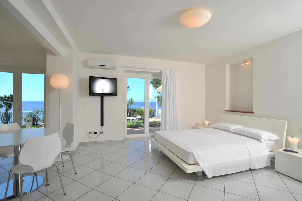 A modern hotel room with a bed, a TV, a dining area with chairs, and a view of the sea through large windows.