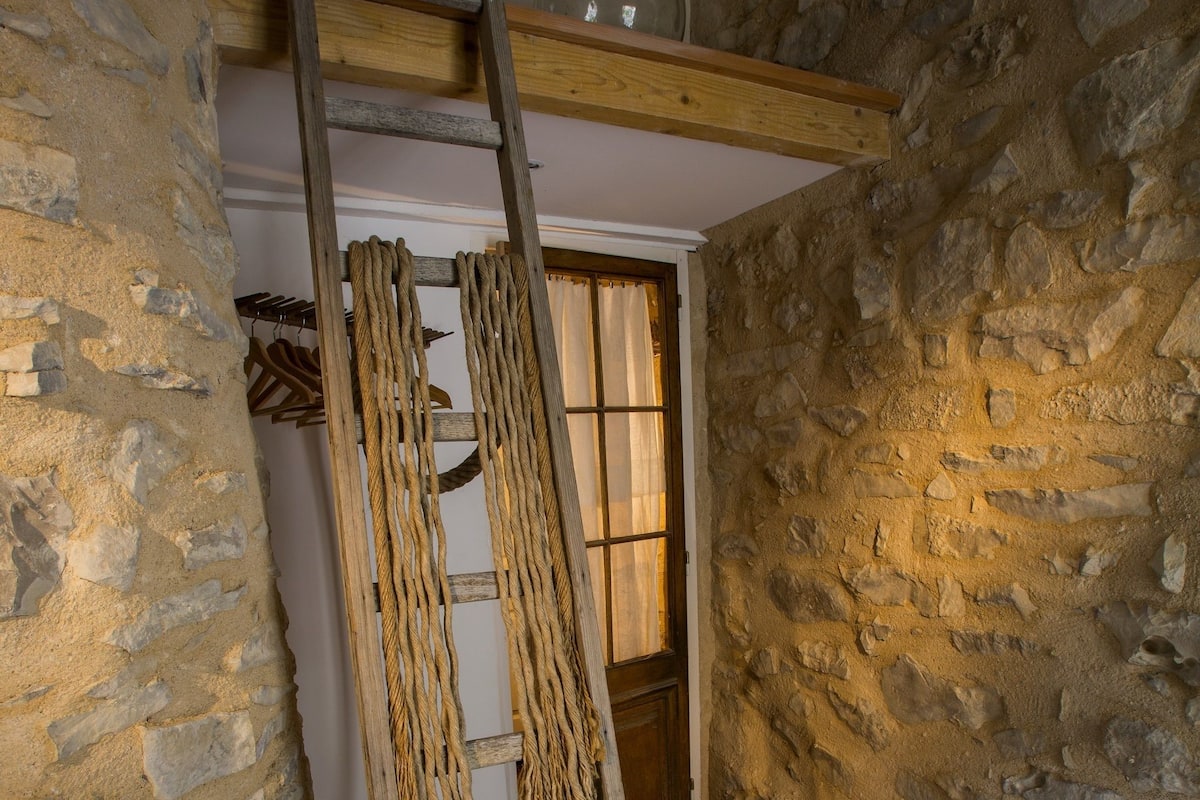 Une pièce rustique aux murs de pierre, avec une échelle en bois appuyée contre le mur, une porte en bois et un tapis blanc sur le sol.