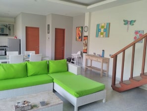 A modern living room with a green sectional sofa, a dining area with a white table and chairs, and a kitchen in the background.
