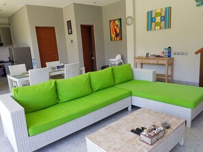 A modern living room with a green sectional sofa, wicker furniture, a wooden coffee table, and a kitchen area in the background.