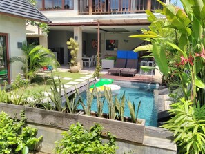 Zona de piscina con una silla de descanso y una mesa de comedor, rodeada de plantas tropicales y un césped cuidado.