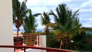A balcony with a view of a swimming pool, palm trees, and a distant view of the ocean.