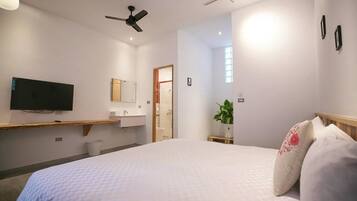 A hotel room with a bed, a TV mounted on the wall, a ceiling fan, a plant, and a wooden bench.