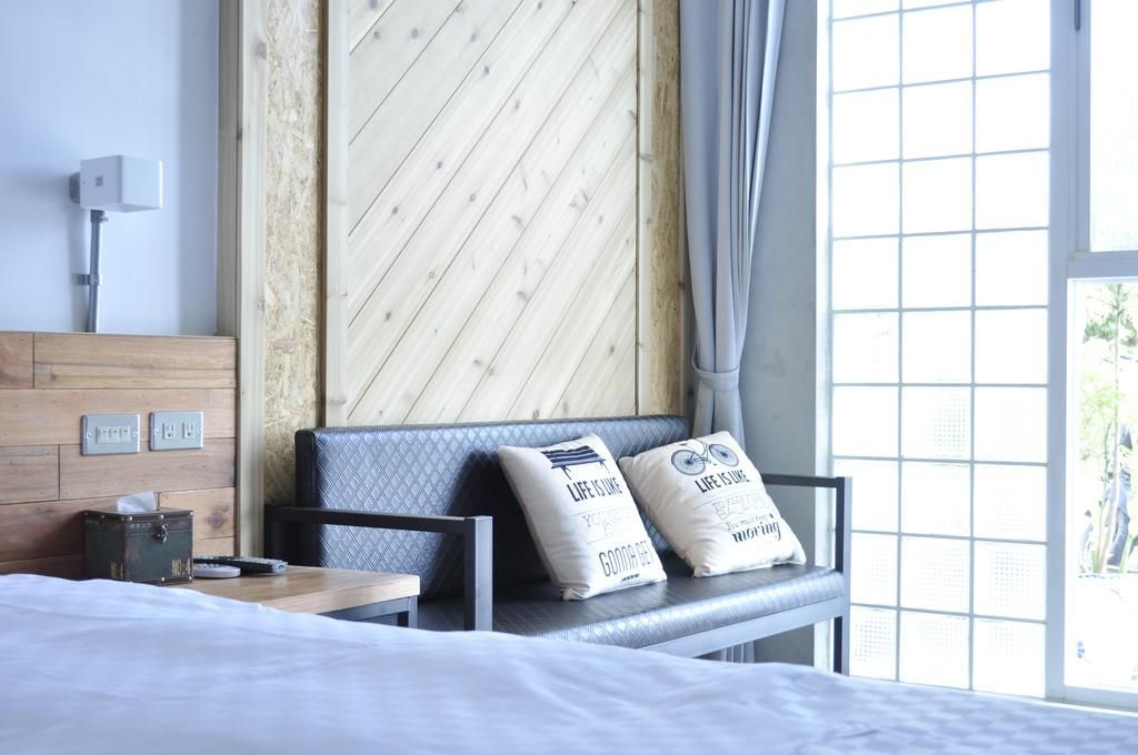Une chambre d’hôtel avec un lit, une banquette avec des coussins, une tête de lit en bois, une table de chevet et une fenêtre donnant sur l’extérieur.