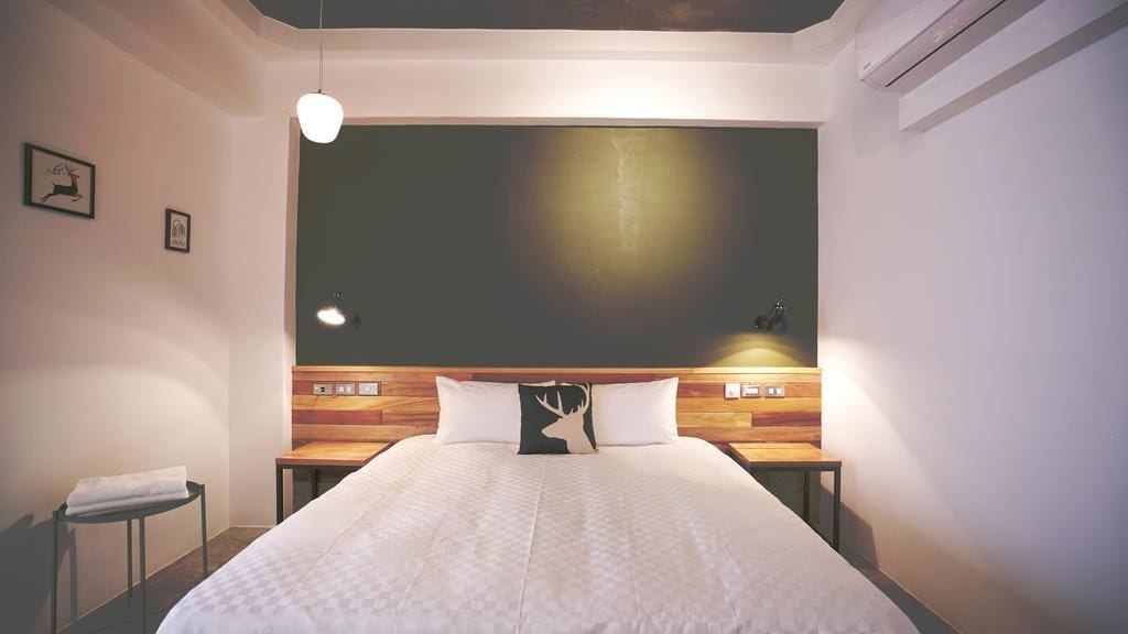 A modern hotel room with a large bed, wooden headboard, and a wall-mounted light fixture.