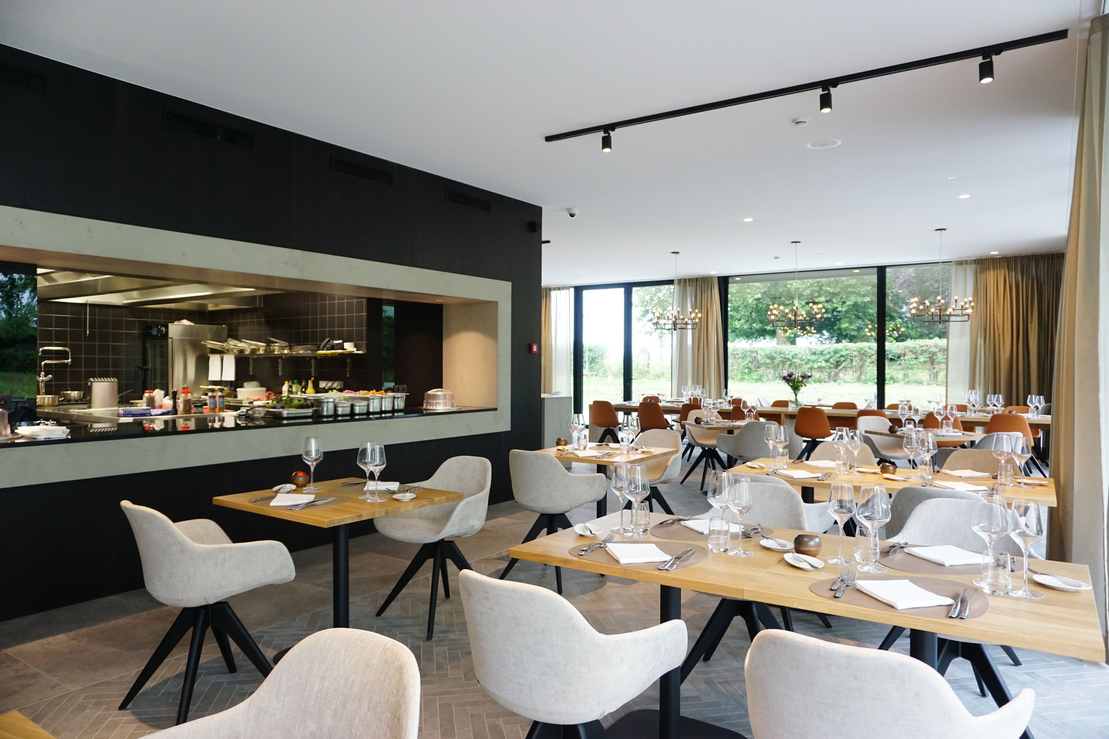 Un comedor moderno con mesas largas, sillas blancas y vistas a la cocina a través de un amplio ventanal.