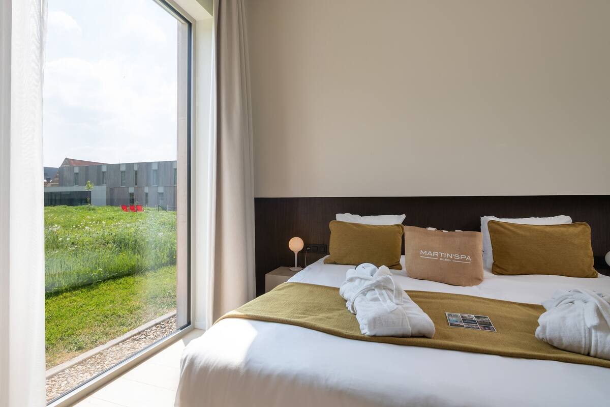 Une chambre d’hôtel avec une grande fenêtre, un lit doté d’une tête de lit marron, un sac de la marque Martin’s Spa, et une vue sur un immeuble et la verdure environnante.