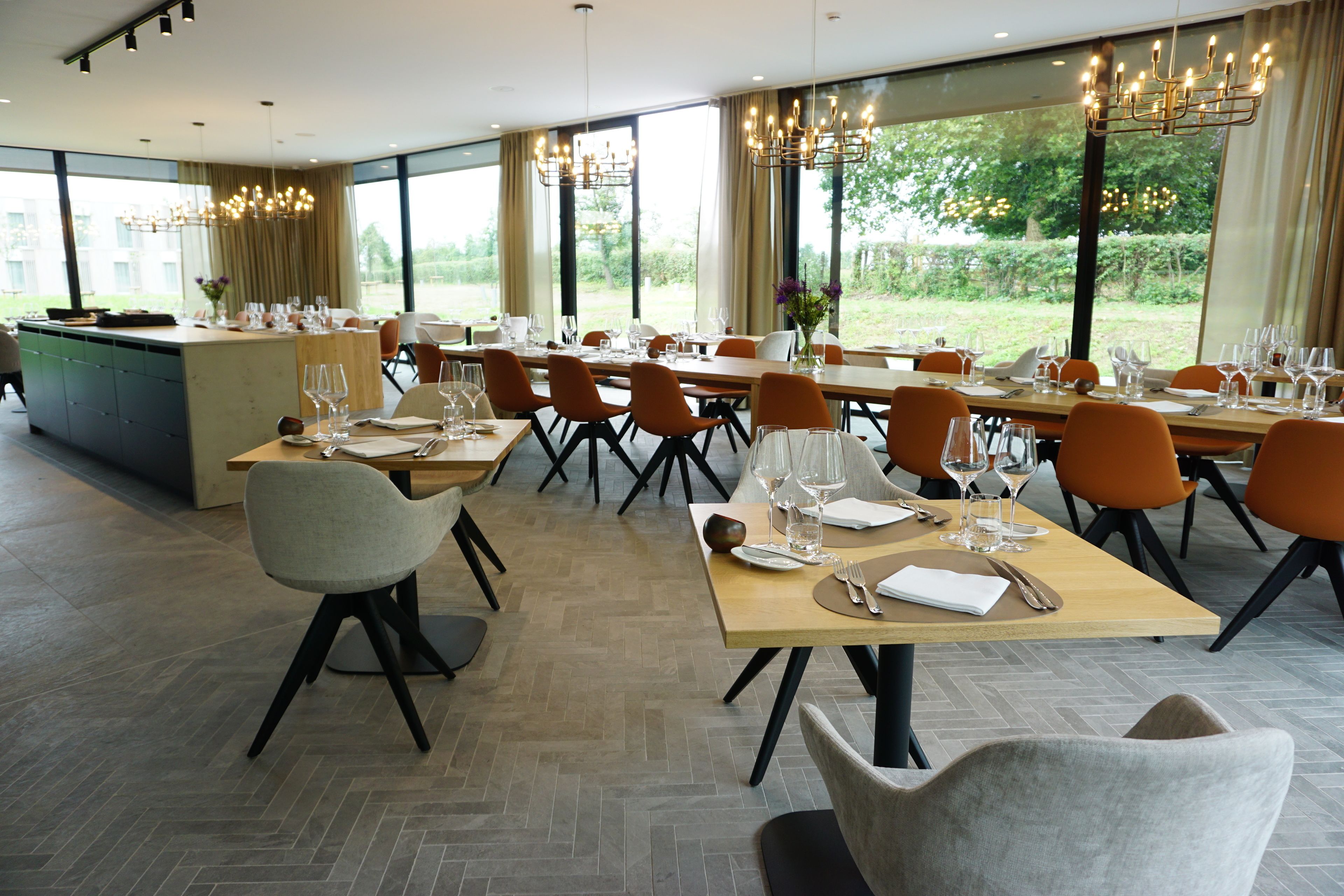 Un vaste espace dédié à la salle à manger, avec de nombreuses tables dressées pour le repas, de grandes fenêtres offrant une vue sur l’extérieur et des lustres modernes.