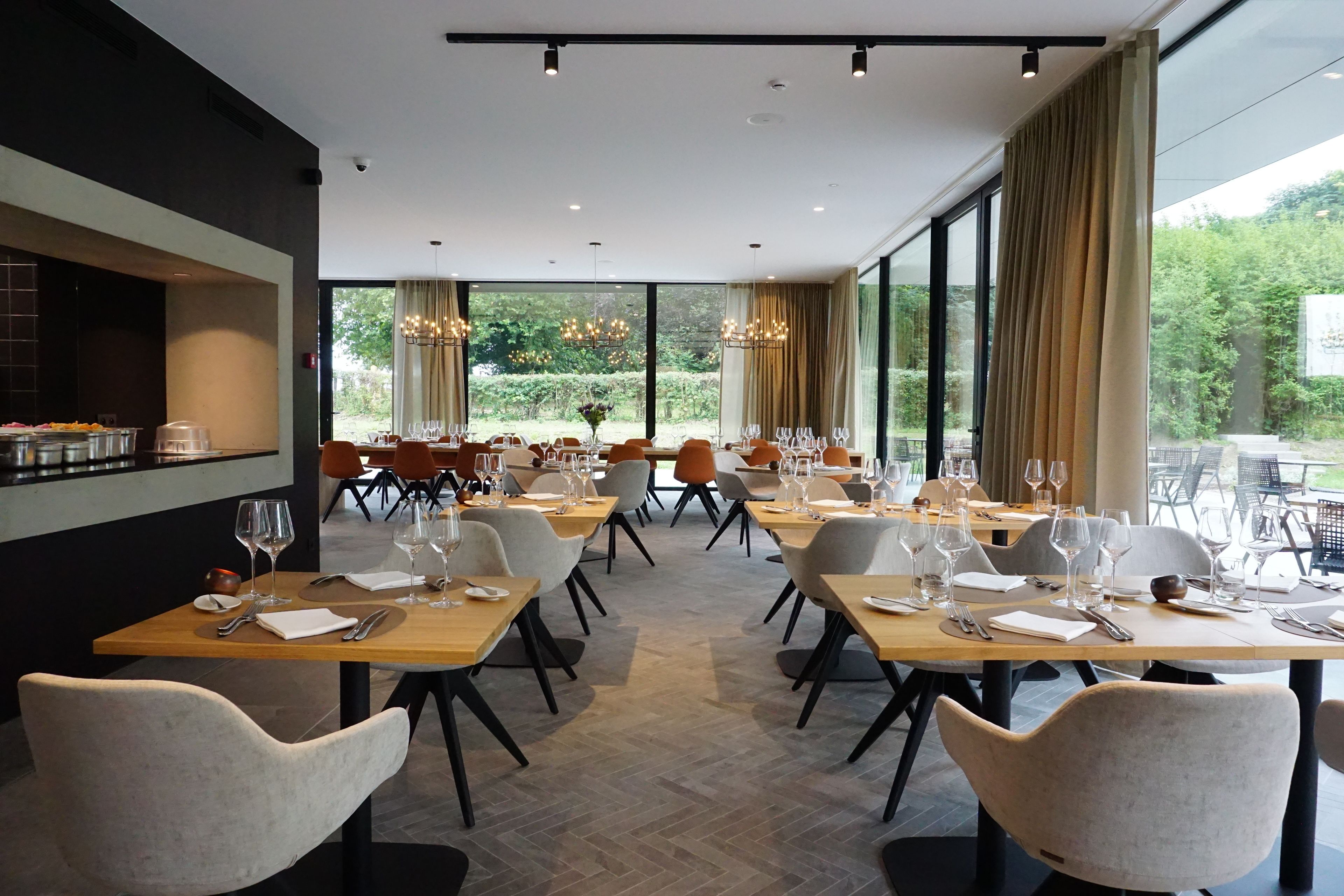 A modern dining area with multiple tables and chairs, large windows offering a view of greenery, and a kitchen area with a counter visible on the left.