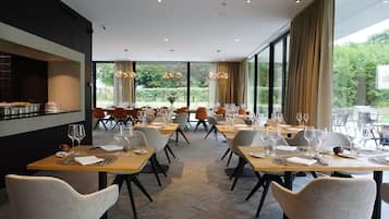 Een moderne eetruimte met meerdere tafels en stoelen, grote ramen met uitzicht op groen, en een keuken met een aanrecht dat links zichtbaar is.