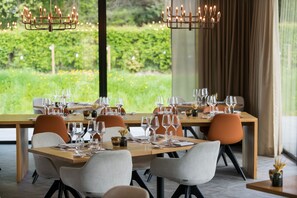 A restaurant with a long table set for dining, surrounded by chairs, and a view of greenery outside.