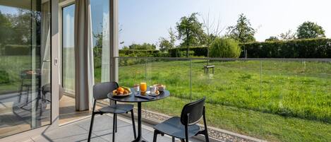 A modern outdoor dining area with a glass partition, two chairs, a table with breakfast items, and a view of a grassy garden.
