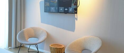 A modern interior with a mounted TV displaying a welcome message, two contemporary chairs, and a wooden stool.