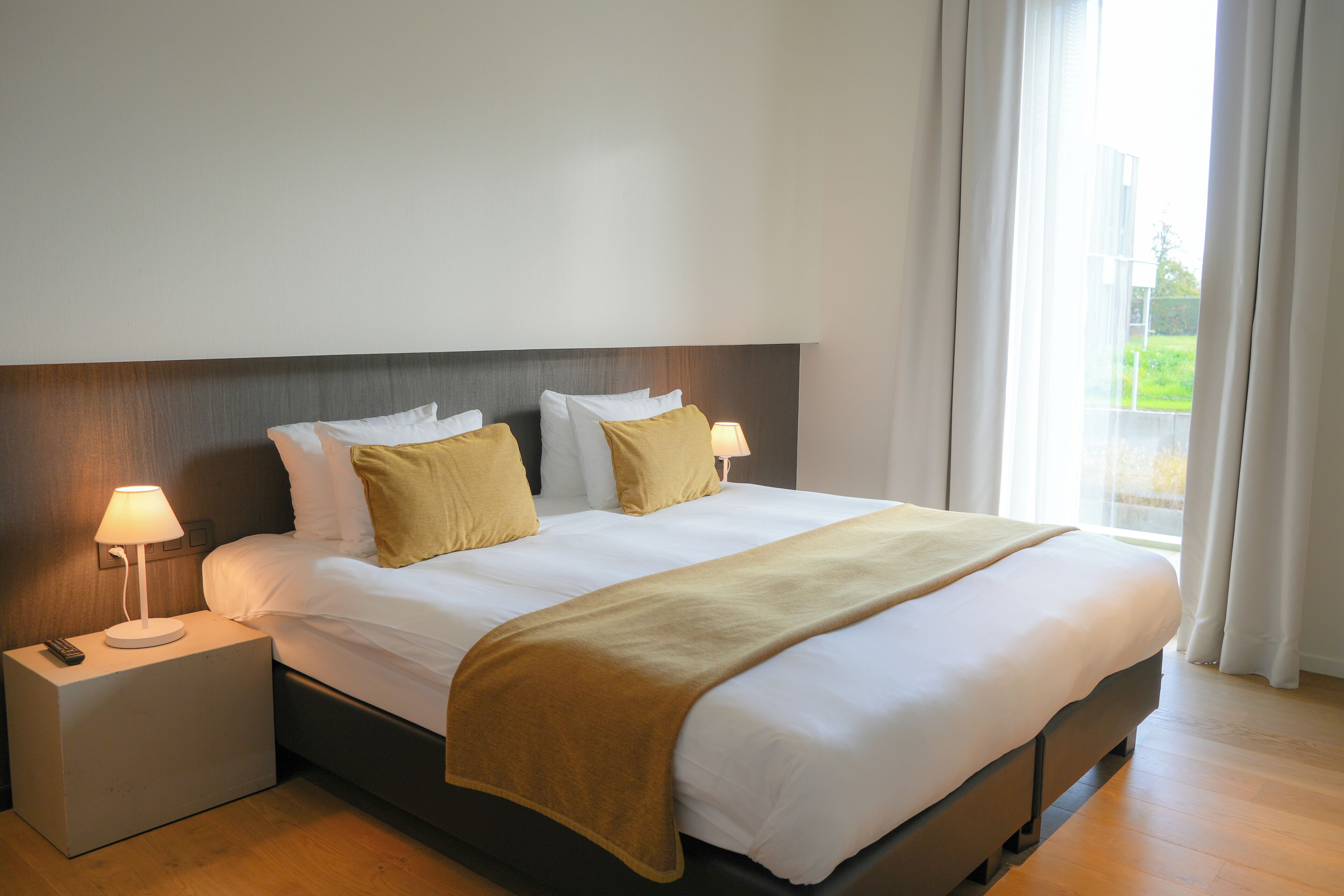 Una cama bien tendida con cabecera, dos lámparas de noche y una ventana con cortinas.