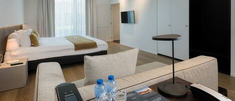 Una habitación de hotel moderna con una cama, un sofá, una mesa de centro con un control remoto, botellas de agua, una revista y un teléfono.