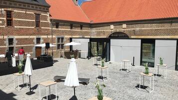 Une cour extérieure dallée de pierres, avec des tables blanches et des parasols, entourée d’immeubles en briques coiffés de toits en tuiles rouges.