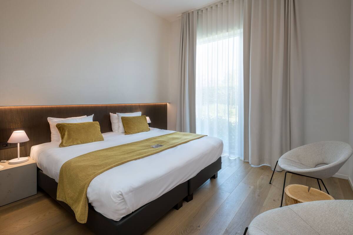 Une chambre d’hôtel moderne avec un grand lit, une lampe de chevet, une chaise et une vue sur l’extérieur à travers des rideaux transparents.