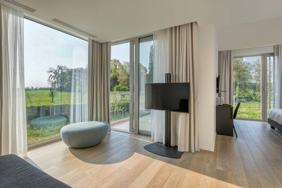 Une chambre d’hôtel moderne dotée d’une grande porte-fenêtre coulissante donnant sur un balcon, d’un téléviseur à écran plat, d’un coin salon confortable et d’un petit bureau.