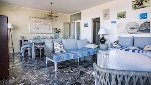 A modern living room with a sofa, a dining table, and clear chairs. There is a chandelier, artwork on the walls, and a lamp. - Helvezia (Jesolo)
