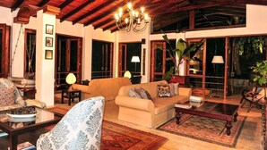 Una sala de estar acogedora con un sofá color beige, sillones estampados y una mesa de centro de madera. Un candelabro cuelga del techo, y hay ventanas amplias con cortinas.